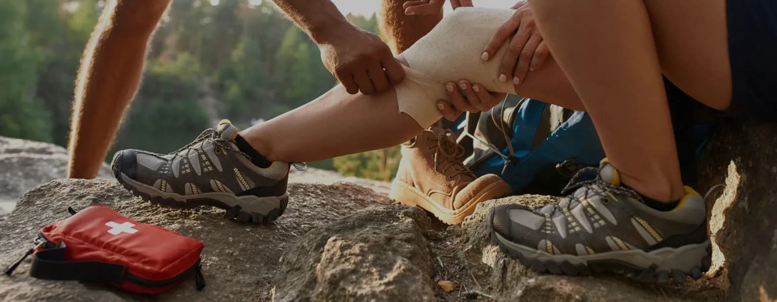 First aid being applied outdoors - someone wrapping a bandage around another person's leg with a red first aid kit visible